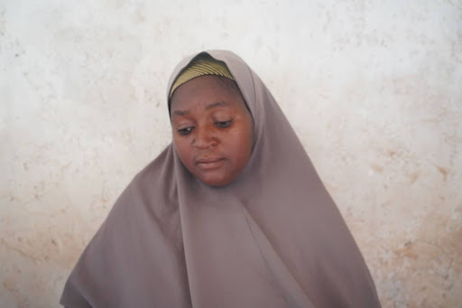 A woman wearing a brown hijab looks down, set against a textured beige wall.