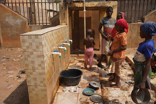 People gathering water from outdoor taps, with bowls and buckets around, in a sunlit area with a tiled wall and wooden structure.