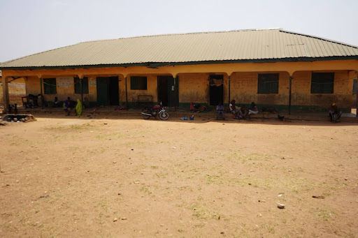 Single-story building with a tin roof, people sitting outside under the shade on a dirt ground, with a motorcycle nearby.