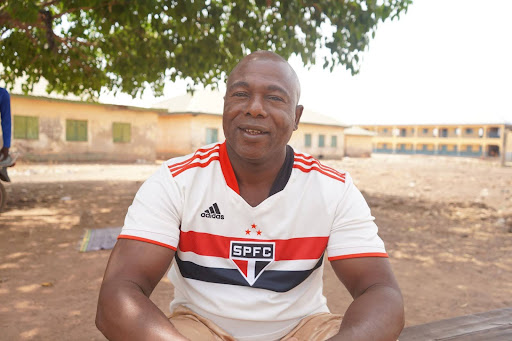 Man sitting outdoors wearing a white SPFC shirt, smiling with trees and buildings in the background.