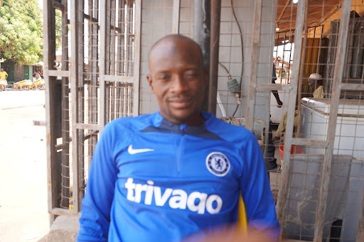 Person wearing a blue Chelsea football jersey sitting outdoors, with a blurred hand in the foreground.