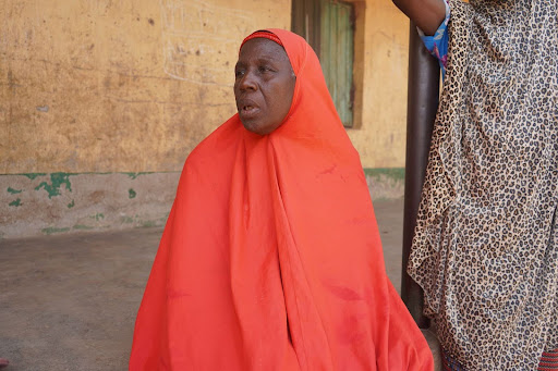 A person in a red garment sits against a yellow wall with a weathered window.