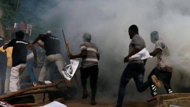 People running through smoke, carrying banners and sticks, in a chaotic scene.