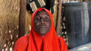 A person wearing a bright red hijab with a nose ring sits against a rustic woven backdrop.