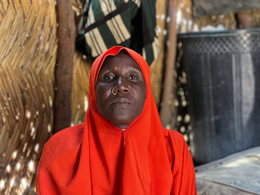 A person wearing a bright red hijab with a nose ring sits against a rustic woven backdrop.