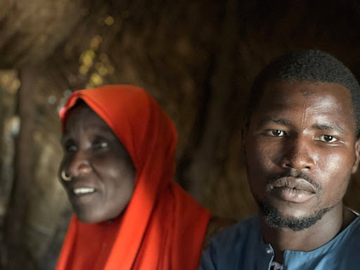 A man and woman sit inside a dimly lit room. The woman wears an orange headscarf, while the man gazes at the camera with a neutral expression.