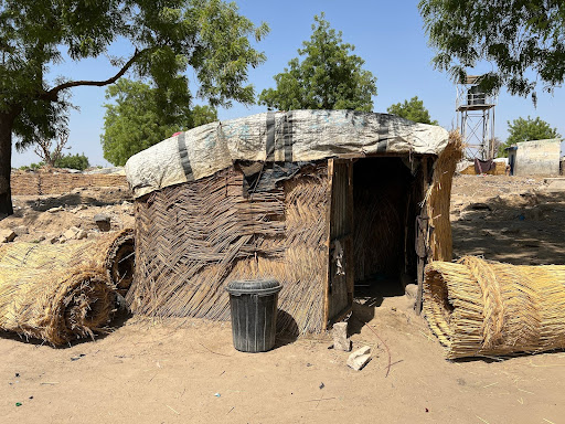 Small hut made of woven straw with rolled mats nearby, set in a sandy area with trees and a distant watchtower.
