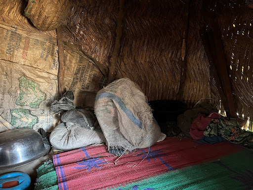 Inside a rustic hut with woven walls, colorful mats, sacks, and a blue container on the floor, creating a simple, cozy atmosphere.
