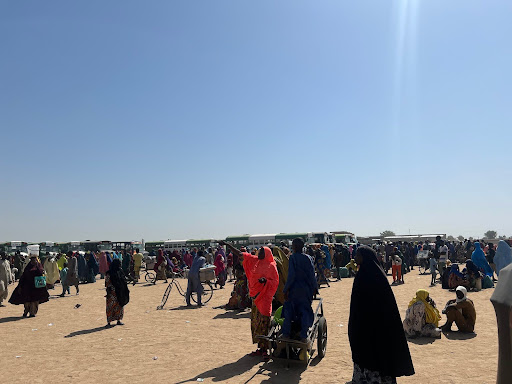 A crowd of people gathered on a sandy field, some sitting and others standing, under a clear blue sky. Buses are lined up in the background.