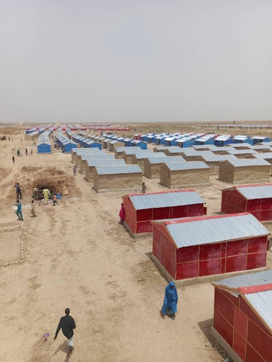 Rows of small shelters with red and blue roofs in a dry, barren landscape. People walk among the structures.