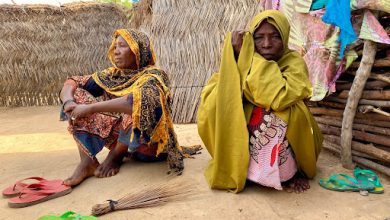 Two women in colorful attire sit outside a straw hut, surrounded by sandals and a straw broom, with a wall of woven material behind them.