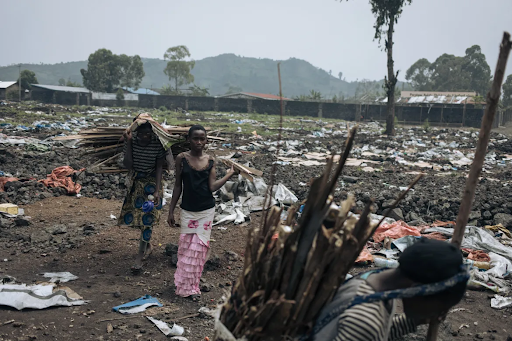 People carrying bundles of sticks walk through a litter-strewn field with trees and mountains in the background.