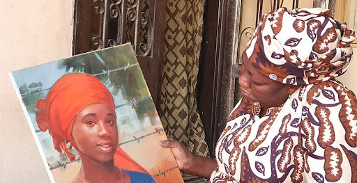 Woman holding a portrait of a young girl in an orange headscarf, both wearing patterned clothing, standing by a doorway.