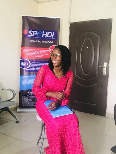 Smiling woman in a pink dress sits holding papers, with a "SPEHDI for Peace and Development" banner and a closed door in the background.
