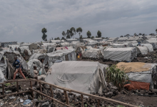 A vast refugee camp with numerous makeshift tents stretches across a rocky landscape, under a cloudy sky, with a few people visible.