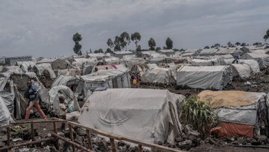 A vast refugee camp with numerous makeshift tents stretches across a rocky landscape, under a cloudy sky, with a few people visible.