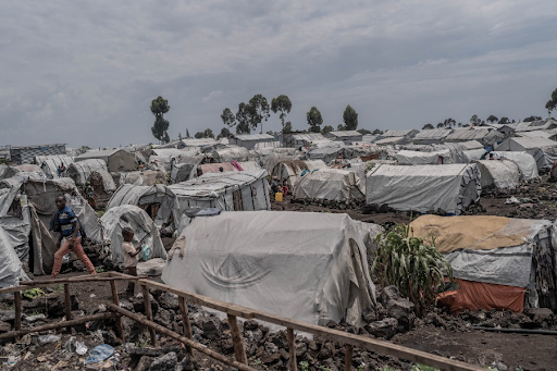 A vast refugee camp with numerous makeshift tents stretches across a rocky landscape, under a cloudy sky, with a few people visible.