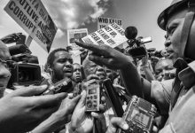 Crowd of reporters interviewing a man, surrounded by protest signs. People hold microphones and cameras in a tense atmosphere. Black and white photo.