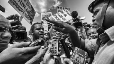 Crowd of reporters interviewing a man, surrounded by protest signs. People hold microphones and cameras in a tense atmosphere. Black and white photo.