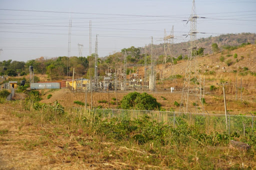 Rural landscape with electrical substation, towers, and fencing amidst dry grass and hills under a clear sky.