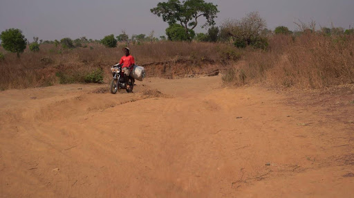 Person riding a motorcycle on a dusty rural path, surrounded by dry grass and sparse trees.