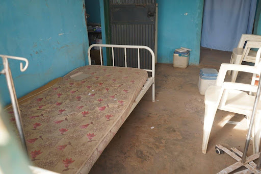 A room with a worn-out bed, plastic chairs, and peeling walls, suggesting a simple, unadorned setting.