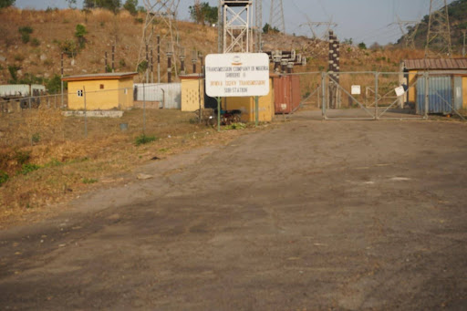 Entrance to a power substation with a sign, buildings, and utility structures surrounded by a fence on a dry, grassy land.