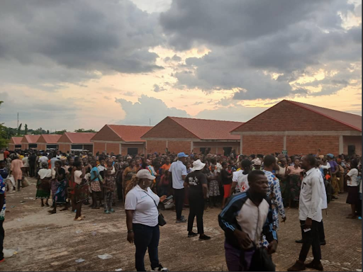 Crowd gathered outdoors near red-roofed buildings at sunset, under a cloudy sky.