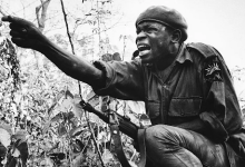 Soldier with a beret, crouched in dense vegetation, points forward with intensity, his rifle slung across his back.