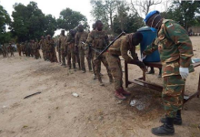 Soldiers line up outdoors to wash hands at a water station, supervised by a peacekeeper wearing a blue helmet.