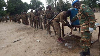 Soldiers line up outdoors to wash hands at a water station, supervised by a peacekeeper wearing a blue helmet.