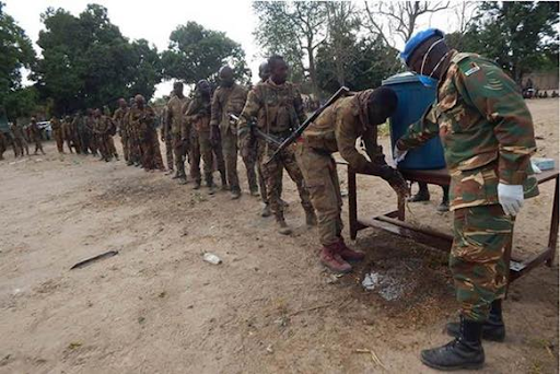 Soldiers line up outdoors to wash hands at a water station, supervised by a peacekeeper wearing a blue helmet.