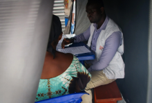 A healthcare worker in a white vest consults with a person seated across a table inside a tent, with papers on the table.
