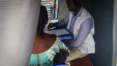 A healthcare worker in a white vest consults with a person seated across a table inside a tent, with papers on the table.