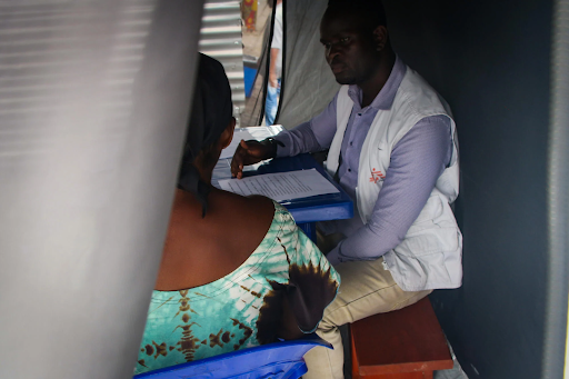 A healthcare worker in a white vest consults with a person seated across a table inside a tent, with papers on the table.