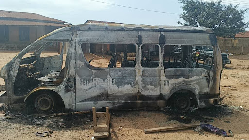 Burned-out minivan in a dirt lot, surrounded by debris, with buildings and a tree in the background.