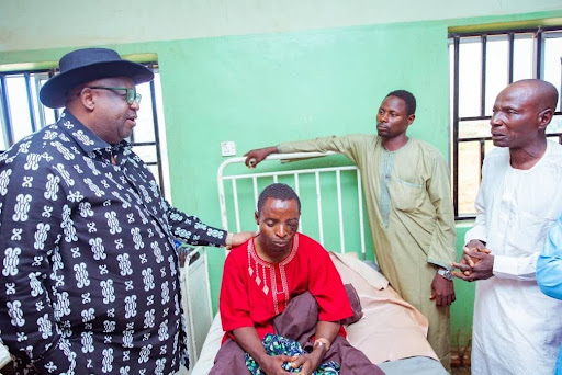 Four men in a hospital room, one patient in bed with others standing around, engaged in conversation, with a supportive gesture.