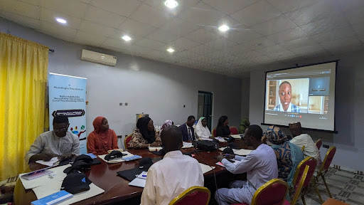 People sitting around a table in a meeting room, watching a virtual presentation on a screen.