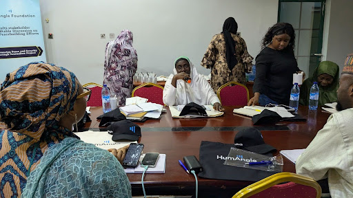 People sitting and standing around a table, engaged in a meeting or discussion, with papers and bottles on the table.