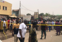 Crowd gathered in a street enclosed by yellow tape, with buildings and signs in the background.