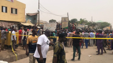 Crowd gathered in a street enclosed by yellow tape, with buildings and signs in the background.