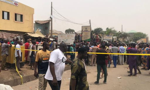 Crowd gathered in a street enclosed by yellow tape, with buildings and signs in the background.