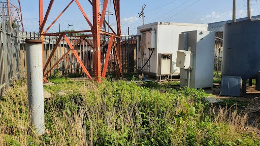 Cell tower site with metal structures and equipment, surrounded by a fence and overgrown grass, under a clear sky.