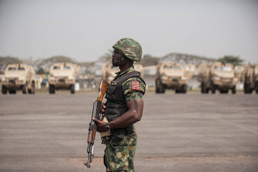 A soldier in camouflage uniform stands guard with a rifle. Armored vehicles are lined up in the background.