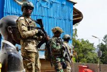Soldiers in camouflage gear stand guard near a blue corrugated metal structure, with a statue and greenery in the background.