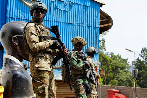 Soldiers in camouflage gear stand guard near a blue corrugated metal structure, with a statue and greenery in the background.