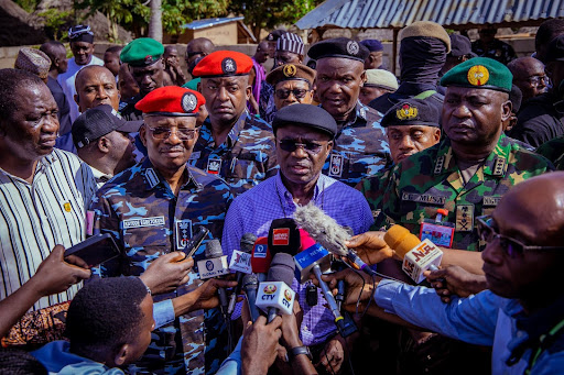A group of officials and military personnel speak to a cluster of reporters and microphones outdoors.