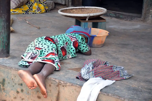 Child in colorful clothing sleeps on a concrete floor, near a plastic bowl and fabric.