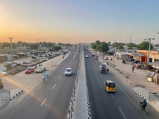 Wide road with vehicles and pedestrians under a clear sky, lined by buildings and trees, during sunset.