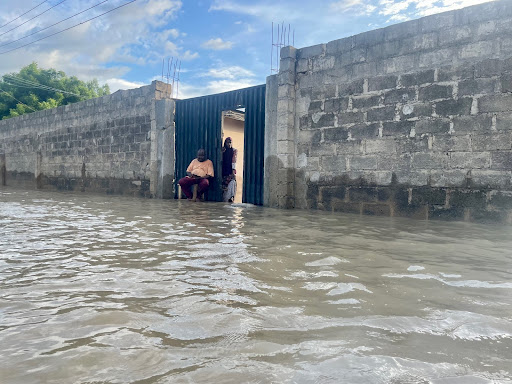 Man sitting at a doorway amid floodwaters, surrounded by a concrete wall and cloudy blue sky above.
